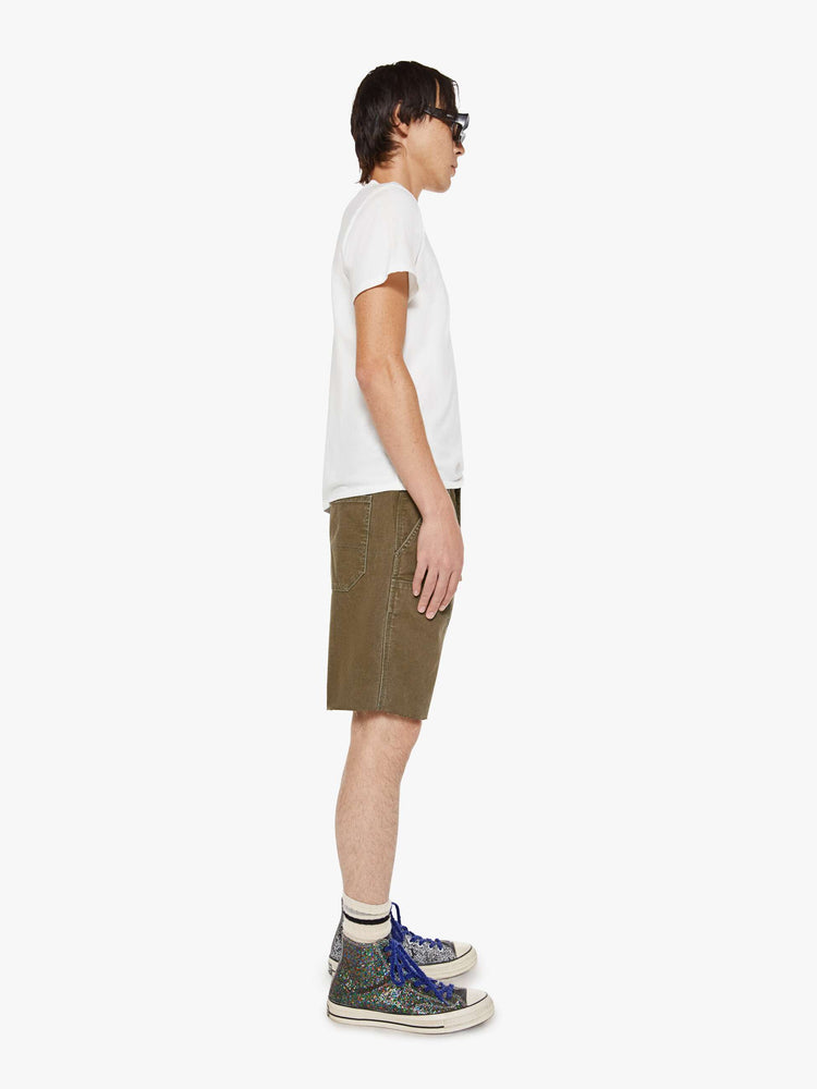 Side view of a man in a dark olive mid-rise shorts with a zip fly, slit pockets, a hammer loop, utility-inspired patches at the knees and a raw, cut-off hem.