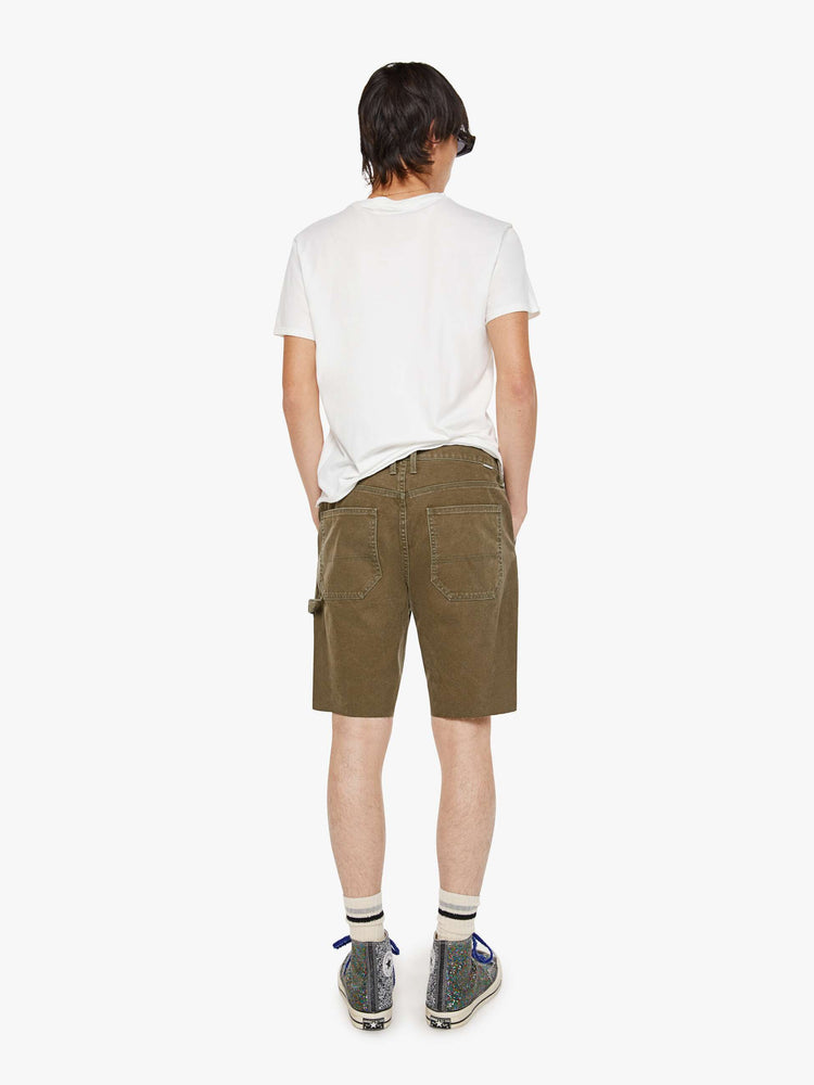 Back view of a man in a dark olive mid-rise shorts with a zip fly, slit pockets, a hammer loop, utility-inspired patches at the knees and a raw, cut-off hem.