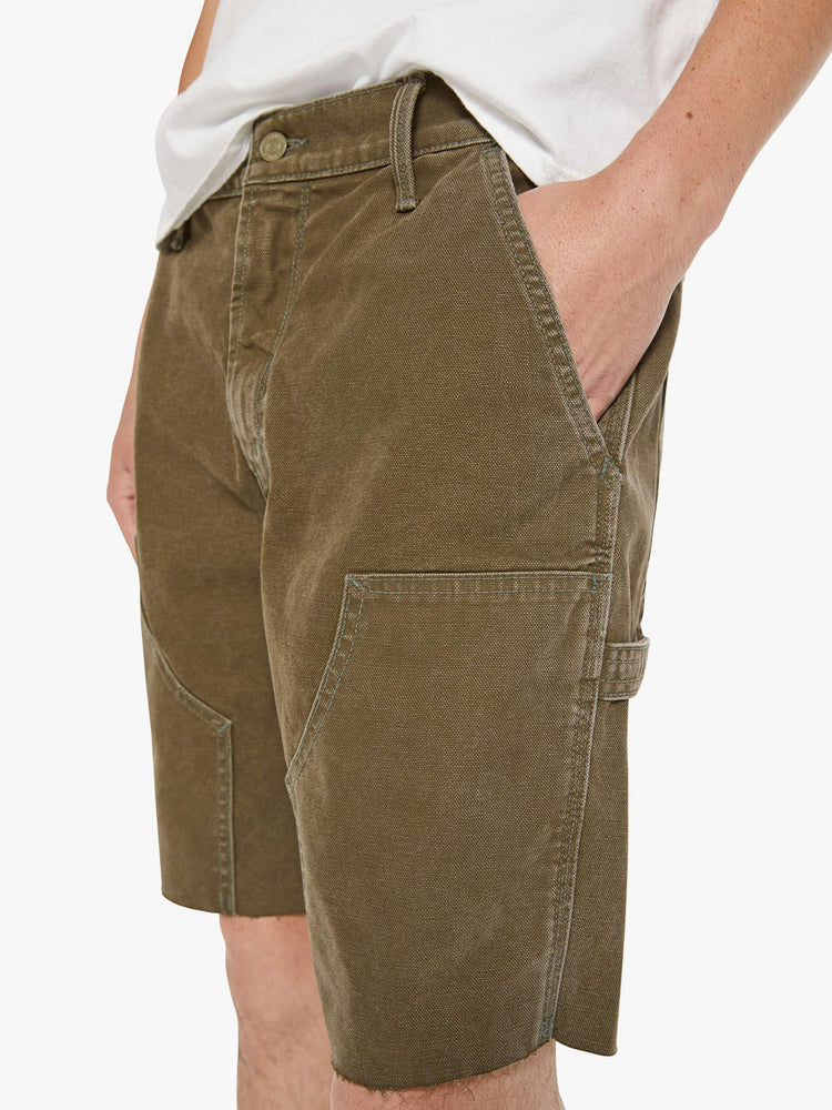 Close up view of a man in a dark olive mid-rise shorts with a zip fly, slit pockets, a hammer loop, utility-inspired patches at the knees and a raw, cut-off hem.