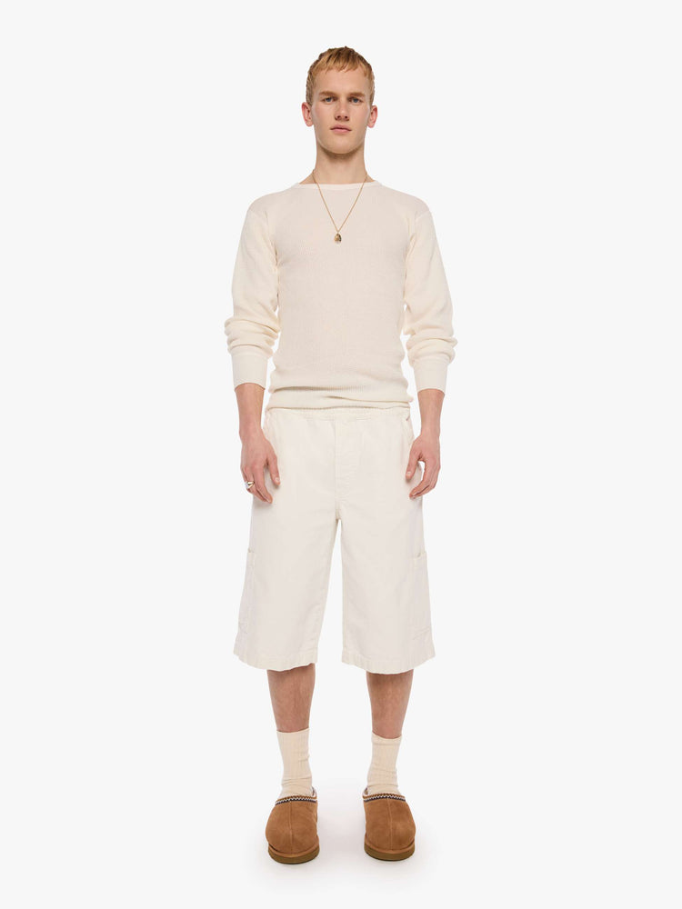 Front view of a man in a cream long shorts with an elastic waistband, faux zip cover, patch pockets on the thighs and a calf-length inseam. 