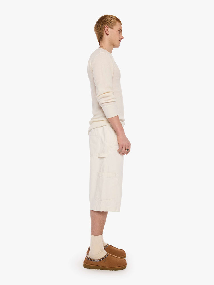 Side view of a man in a cream long shorts with an elastic waistband, faux zip cover, patch pockets on the thighs and a calf-length inseam. 