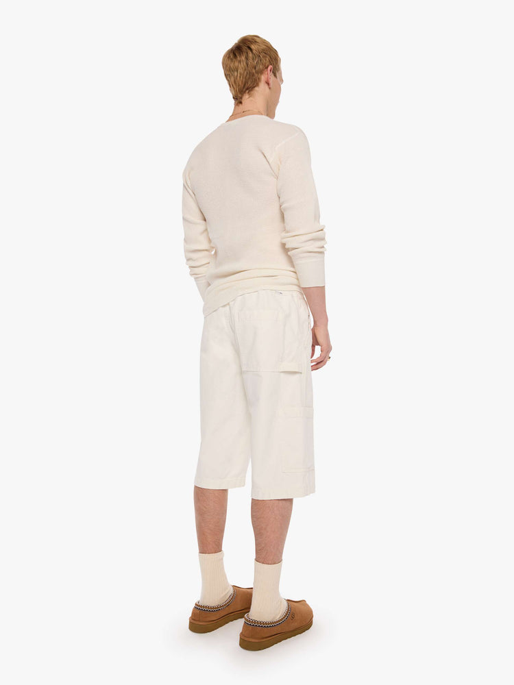 Back view of a man in a cream long shorts with an elastic waistband, faux zip cover, patch pockets on the thighs and a calf-length inseam. 