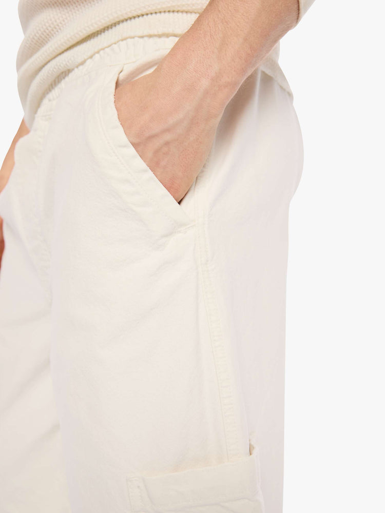 Close up view of a man in a cream long shorts with an elastic waistband, faux zip cover, patch pockets on the thighs and a calf-length inseam. 