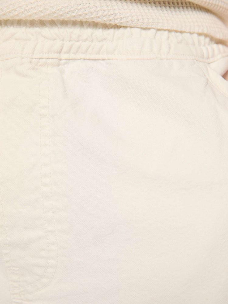 Close up view of a man in a cream long shorts with an elastic waistband, faux zip cover, patch pockets on the thighs and a calf-length inseam. 