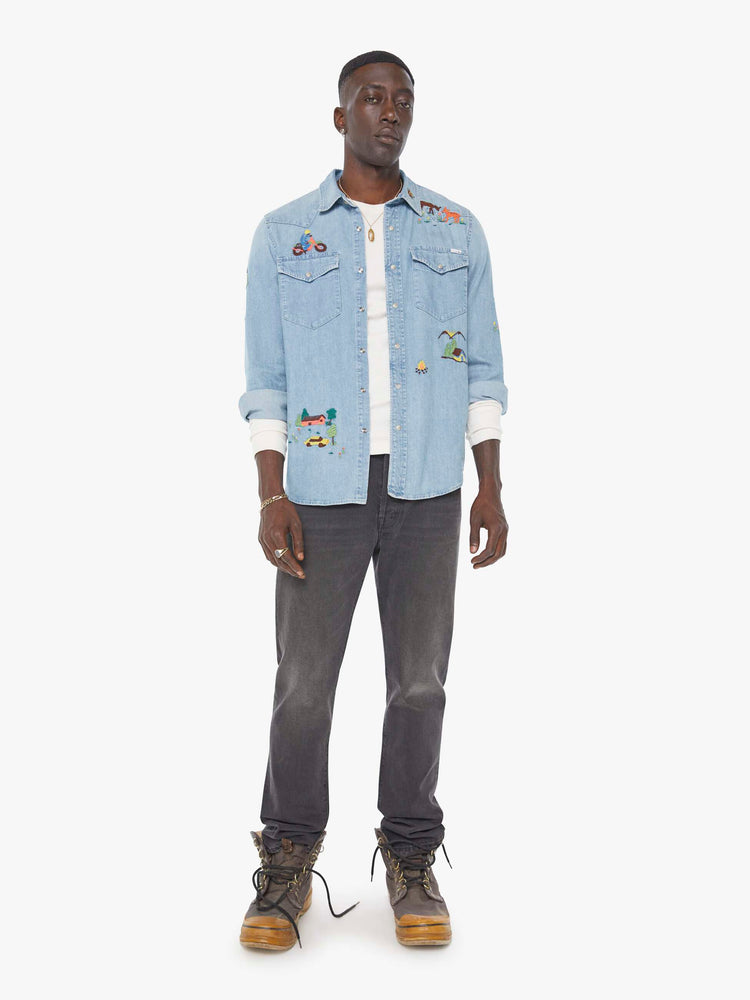Full body view of a man in a light blue denim button up with fading and colorful embroidery throughout.