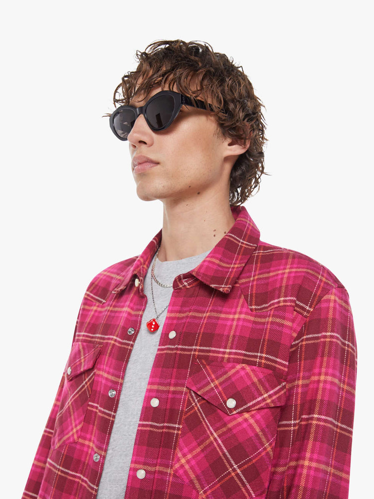 Close up view of a man in a hot pink and red plaid print classic button down shirt with flap patch pockets, long sleeves and a curved hem.