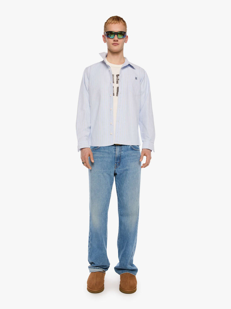 Full body view of a man in light blue shirt with off white and black stripes and a navy M embroidered on the chest.