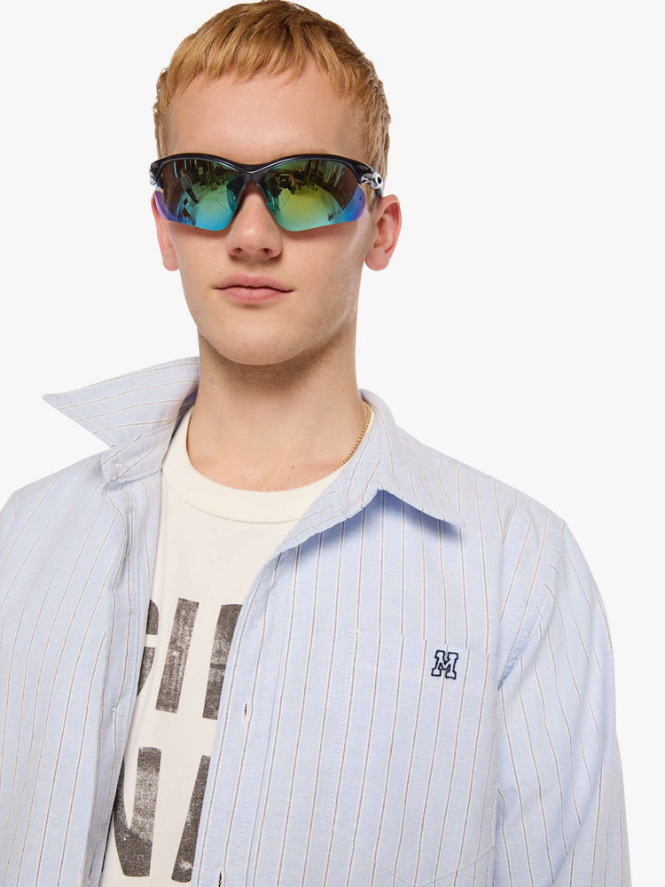 Close up view of a man in light blue shirt with off white and black stripes and a navy M embroidered on the chest.