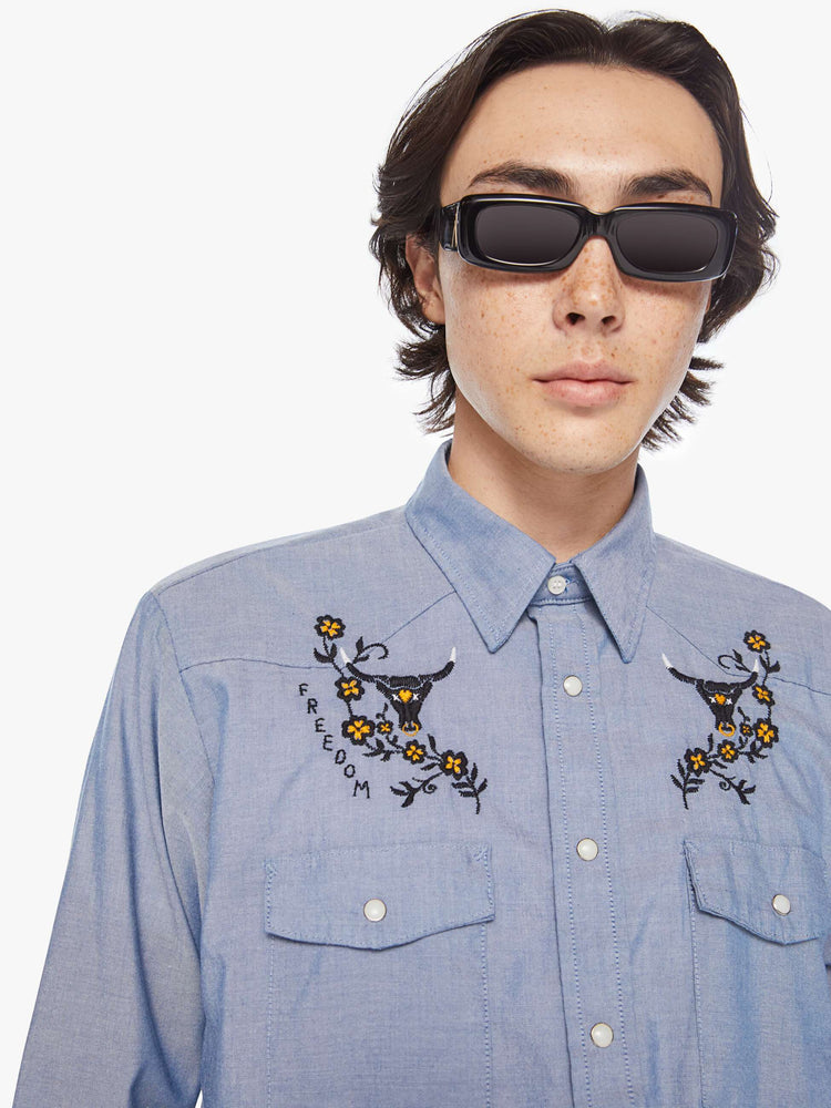 Close up view of a men classic long sleeve button-up with Western-inspired patch pockets in a light blue hue with embroidered bulls, flowers and text on the chest.