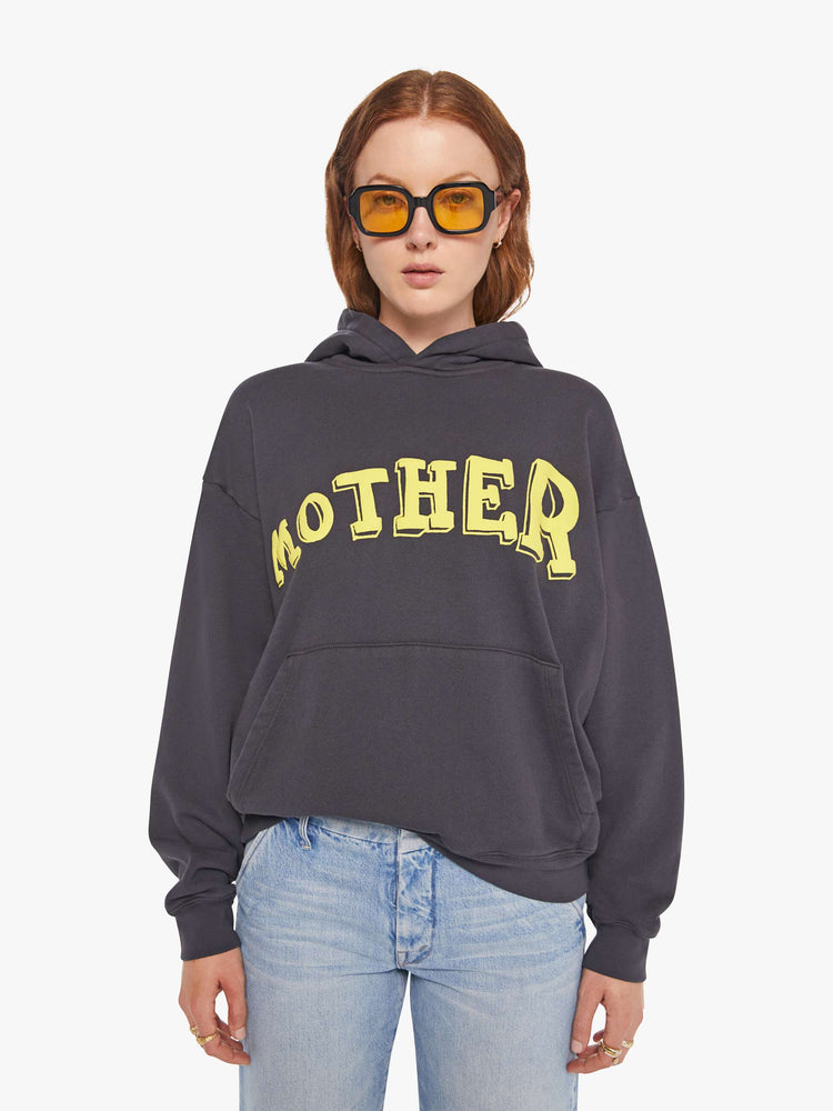 WOMAN front view of an oversized hoodie with drop shoulders, ribbed hems and a front patch pocket in faded black, the hoodie features MOTHER's name in light yellow on the front.