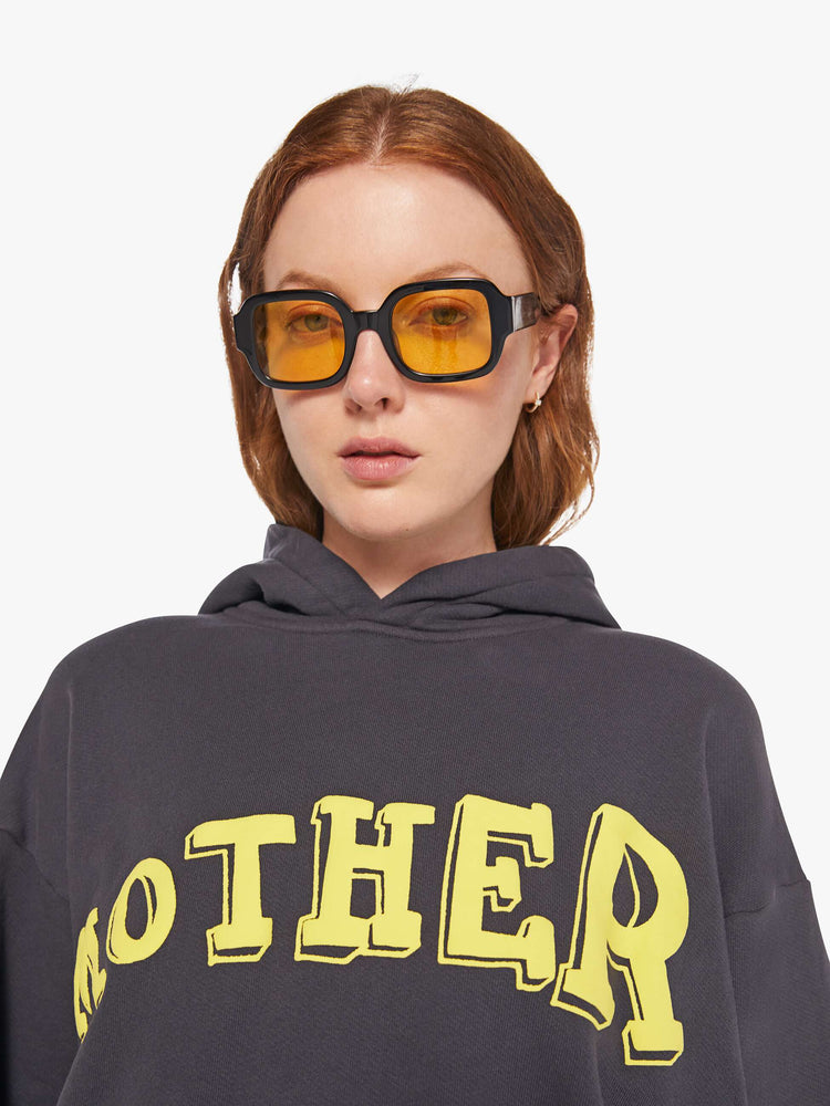WOMAN close up  view of an oversized hoodie with drop shoulders, ribbed hems and a front patch pocket in faded black, the hoodie features MOTHER's name in light yellow on the front.