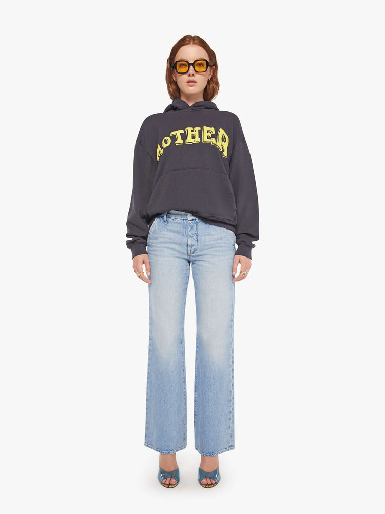 WOMAN full body view of an oversized hoodie with drop shoulders, ribbed hems and a front patch pocket in faded black, the hoodie features MOTHER's name in light yellow on the front.