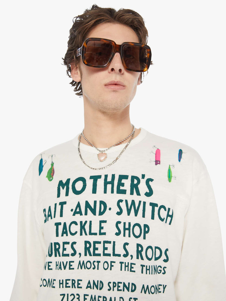 MEN close up view of rugby style tee in off white with a green text graphic on the front and colorful lures across the shoulders.