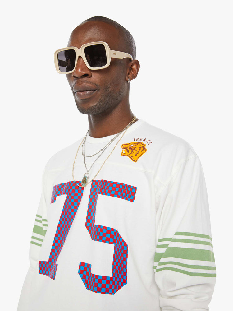 MEN close up view in a white rugby-style tee with a crew neck, long sleeves with a checkered "75" graphic on the front, mint-green stripes on the sleeves and a tiger near the neck.