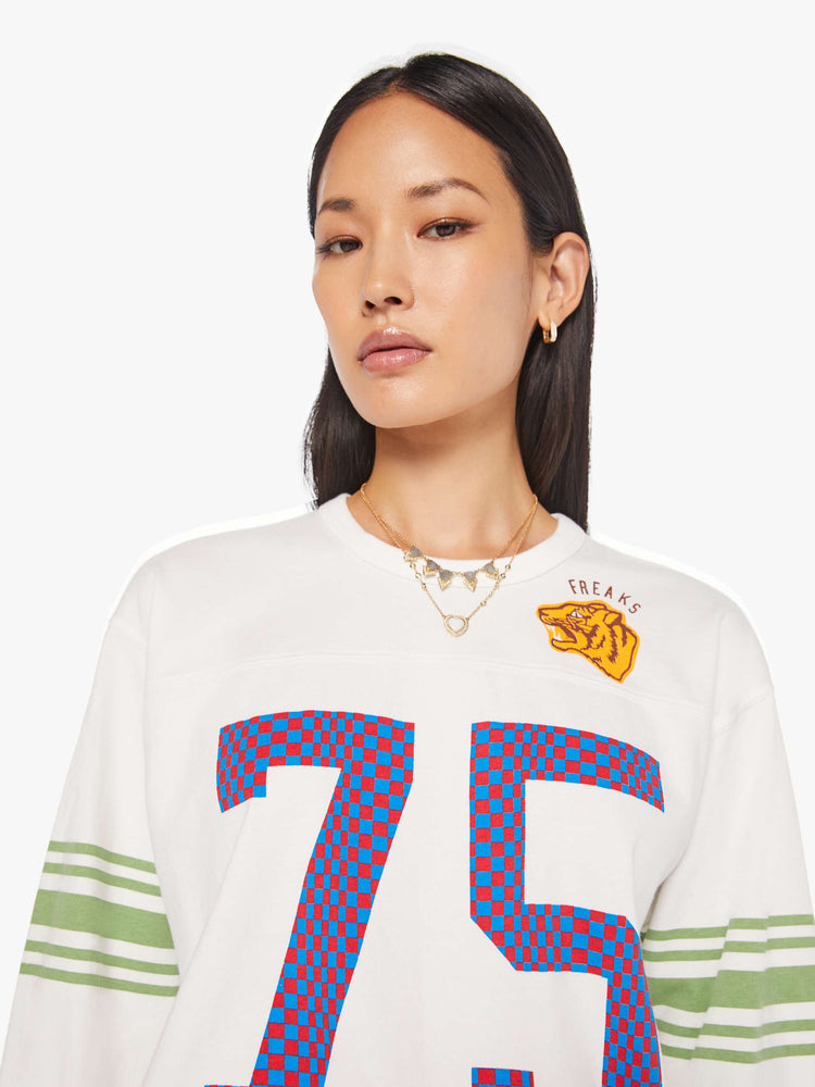 WOMAN close up view in a white rugby-style tee with a crew neck, long sleeves with a checkered "75" graphic on the front, mint-green stripes on the sleeves and a tiger near the neck.