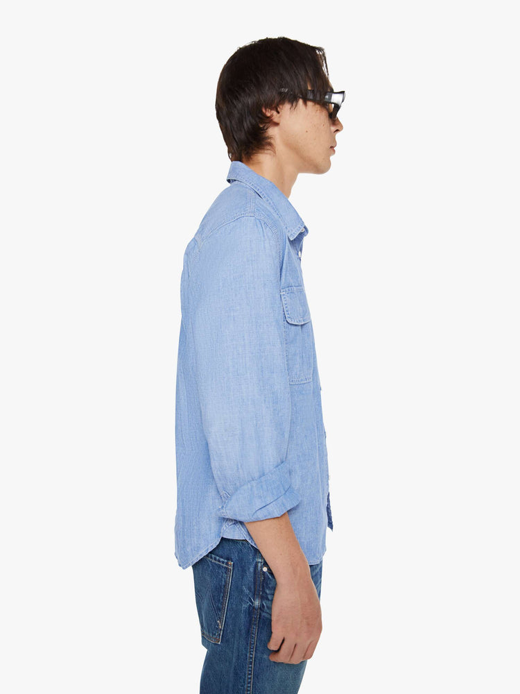 Side view of a man in a light blue denim button-up with patch pockets, long sleeves and a curved hem.