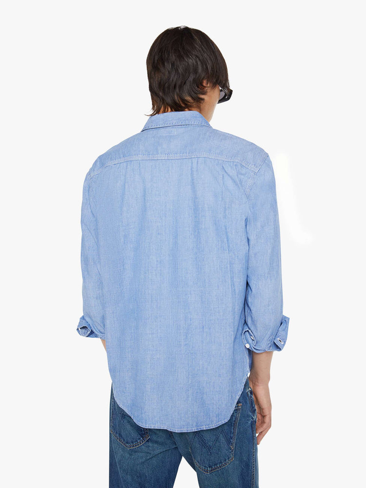 Back view of a man in a light blue denim button-up with patch pockets, long sleeves and a curved hem.