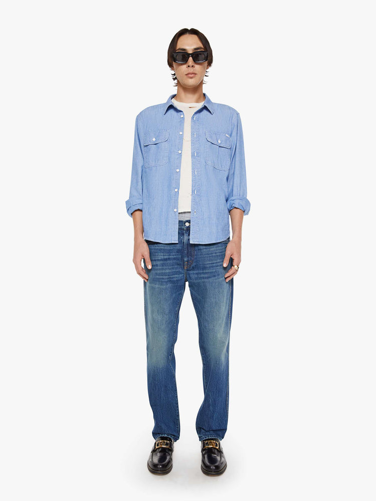 Full body view of a man in a light blue denim button-up with patch pockets, long sleeves and a curved hem.