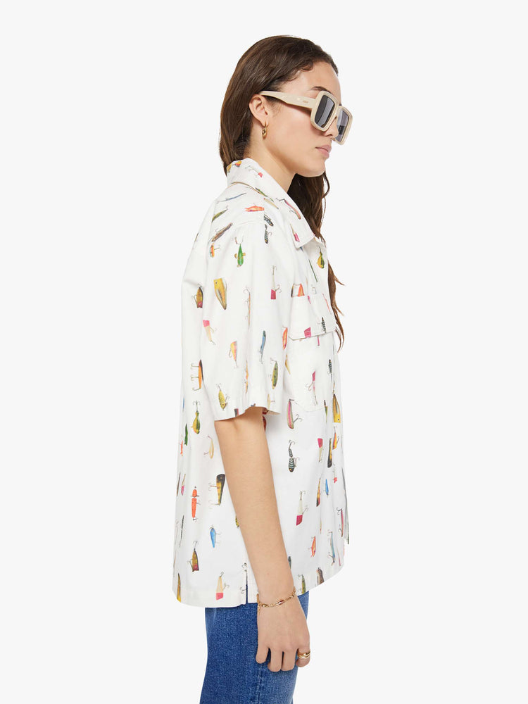 WOMAN side view of a bright white with a colorful fishing lure print short-sleeve button-up with front patch pockets and a boxy fit. 
