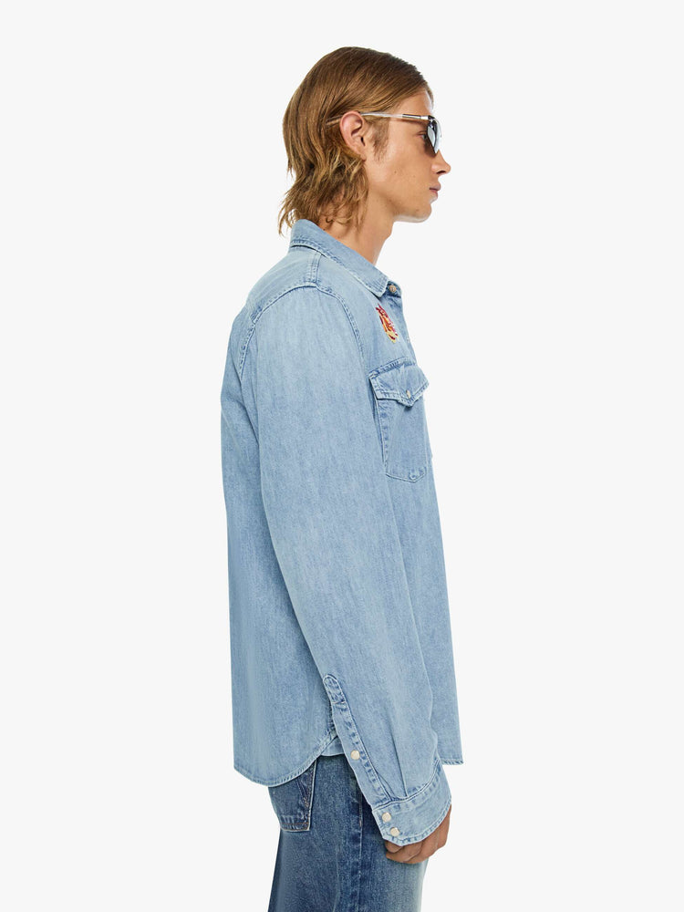 Side view of a man in a light blue with pearl snap buttons, fading at the shoulders and tigers embroidered on the chest classic button-up shirt with front patch pockets, a curved hem and slightly oversized fit. 