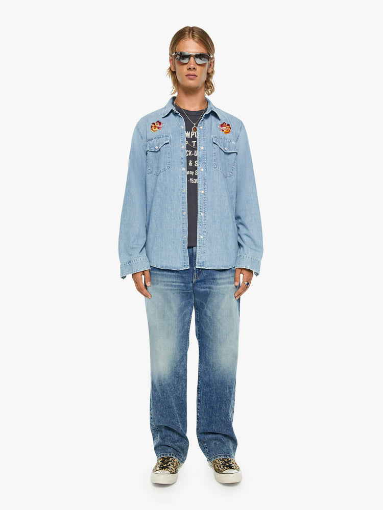 Full body view of a man in a light blue with pearl snap buttons, fading at the shoulders and tigers embroidered on the chest classic button-up shirt with front patch pockets, a curved hem and slightly oversized fit. 