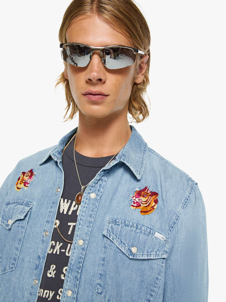 Close up view of a man in a light blue with pearl snap buttons, fading at the shoulders and tigers embroidered on the chest classic button-up shirt with front patch pockets, a curved hem and slightly oversized fit. 