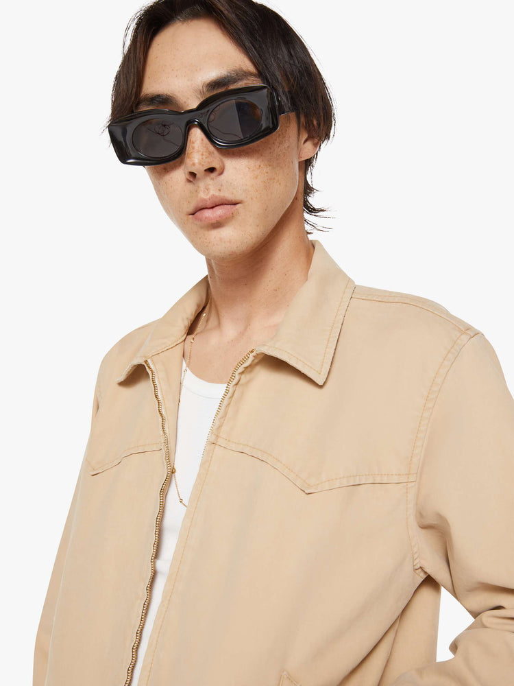 Close up view of a man in a light khaki boxy zip-up rain jacket with a Western-inspired yoke, slit pockets, elastic hems and a slightly cropped fit.