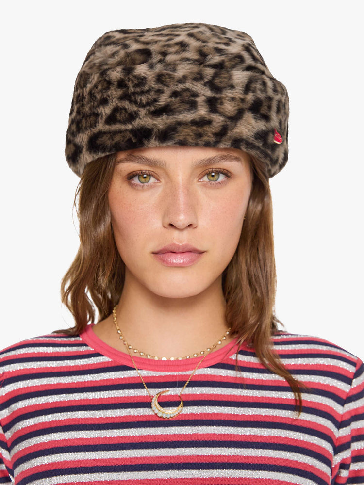 Front view of a woman in a hat with a wide folded brim and a decorative gold-toned pin with a pink heart.