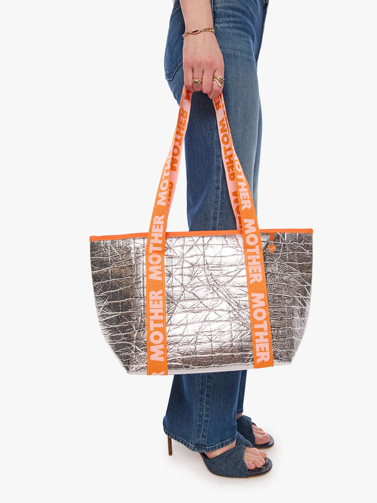 Model carrying metallic silver with a branded orange-and-baby-pink strap and decorative heart charm, and measures 11-inches wide and 10-inches tall with a 5 1/2-inch gusset.