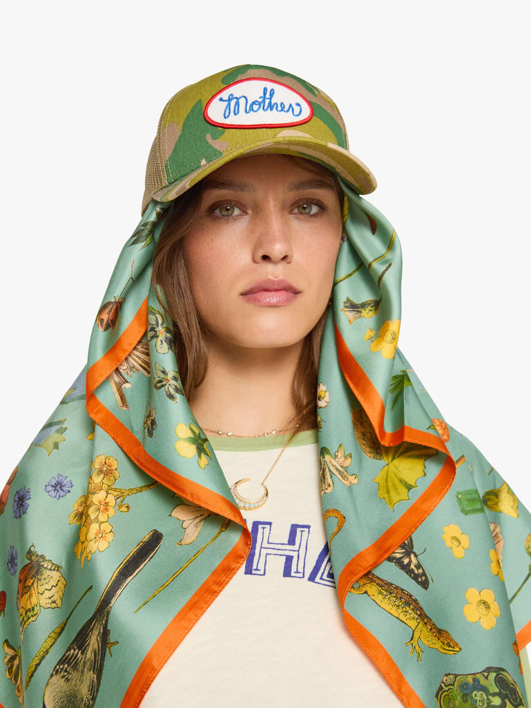 On model wearing a teal-green scarf with oversized printed flowers, gems, and creatures throughout.