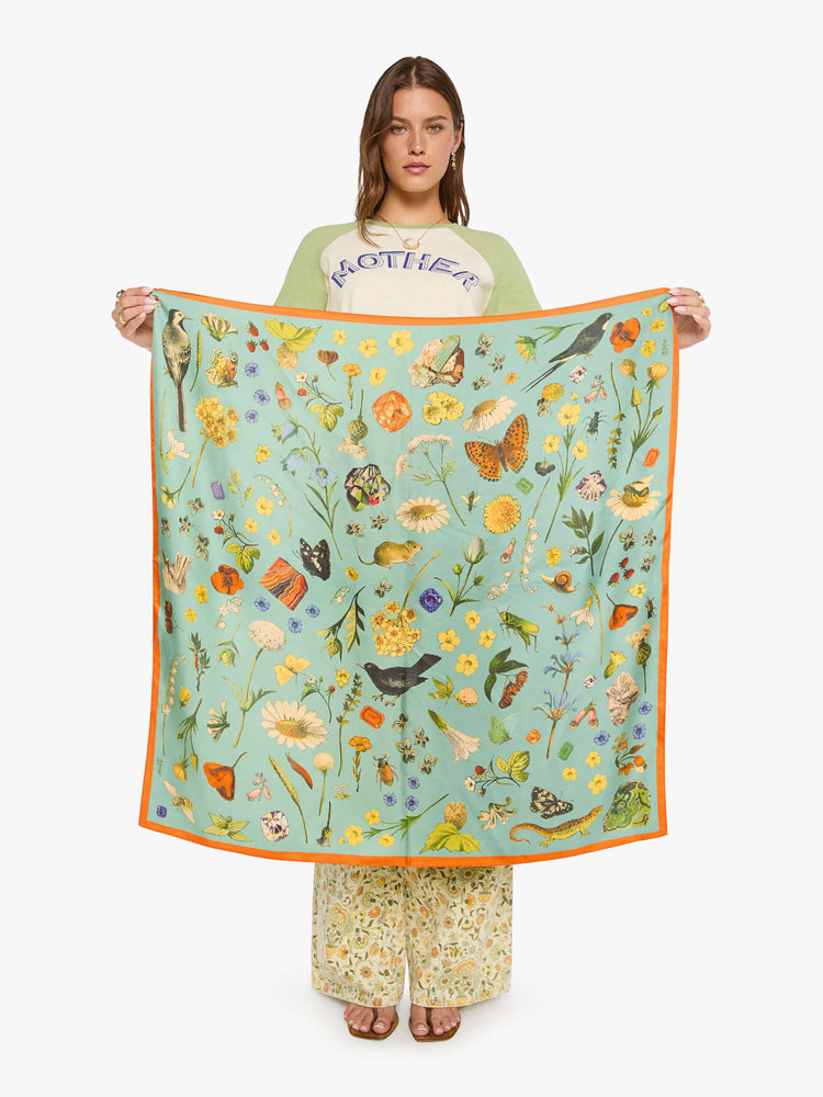 Model holding a teal-green scarf with oversized printed flowers, gems, and creatures throughout.