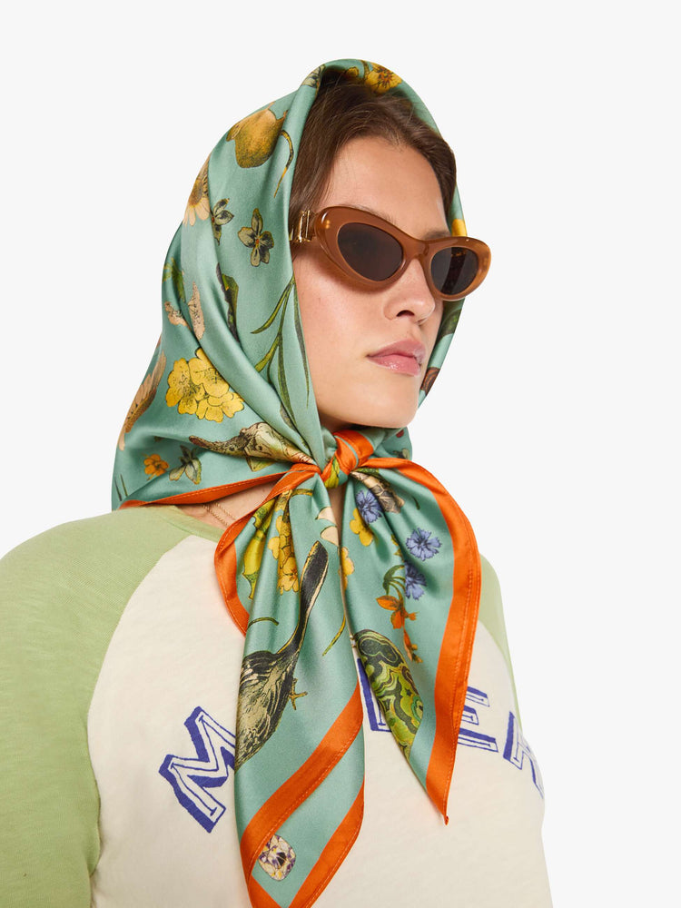 On model wearing view of a teal-green scarf with oversized printed flowers, gems, and creatures throughout.