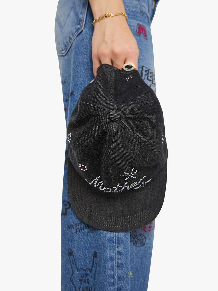 Top view of a woman holding dad hat designed in a faded black 100% cotton denim with MOTHER's name in rhinestones on the front.