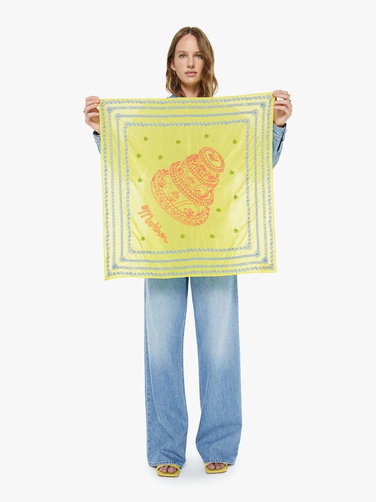 Model holding 26" square scarf made from a cotton blend in pale yellow with piping-inspired embroidery in pink with baby blue trim.