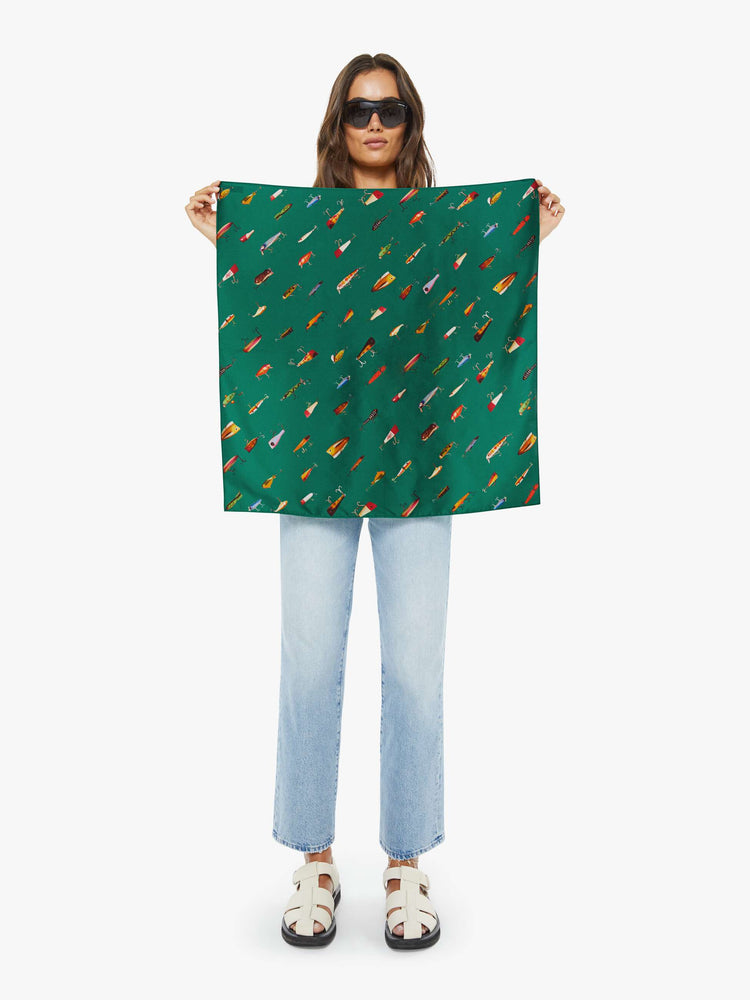 Model holding view of a green-teal fish print  of a 25-inch-wide square scarf designed to be worn around the neck, on your head or tied on a bag. 