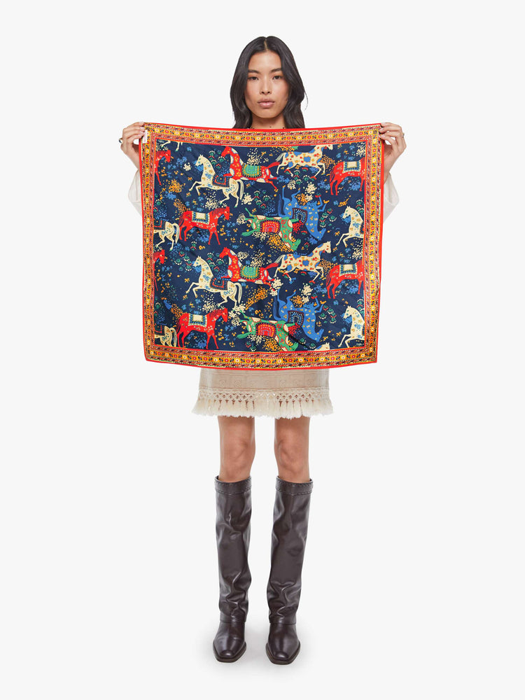 Model holding square scarf made from 100% silk in navy blue with a folk art-inspired horse print and orange trim.