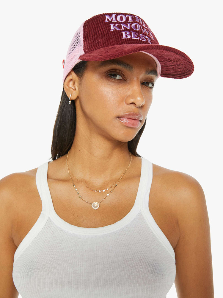 A woman wearing a vintage-inspired trucker hat in a maroon-colored corduroy with embroidered text on the front and pink mesh in the back.