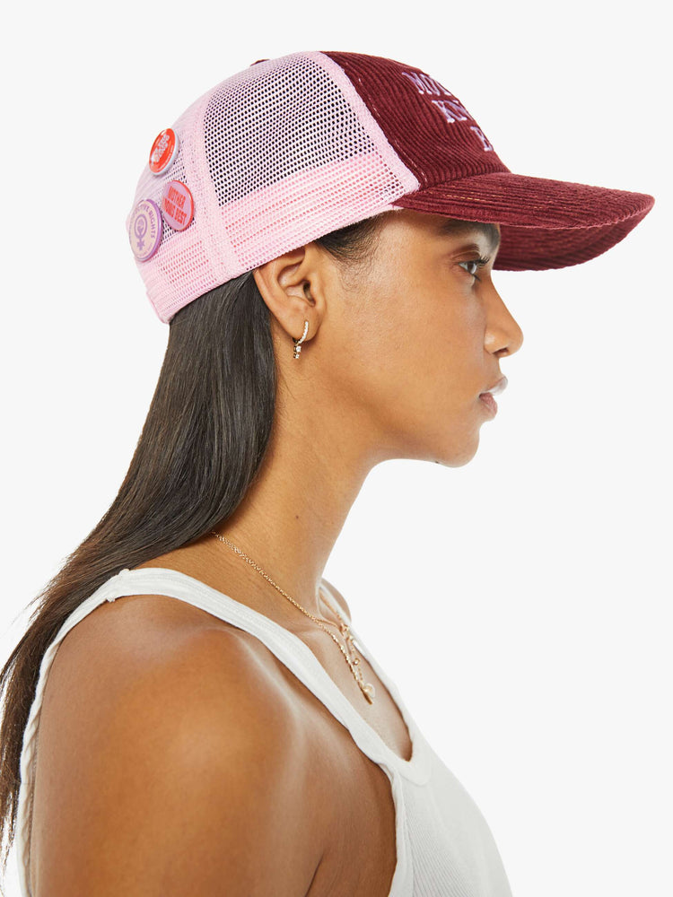 Side view of a woman wearing a vintage-inspired trucker hat in a maroon-colored corduroy with embroidered text on the front, pink mesh in the back and three statement-making pins.