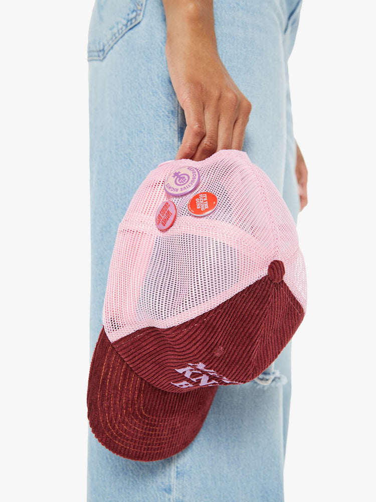 Side view of a woman holding a vintage-inspired trucker hat in a maroon-colored corduroy with embroidered text on the front, pink mesh in the back and three statement-making pins.