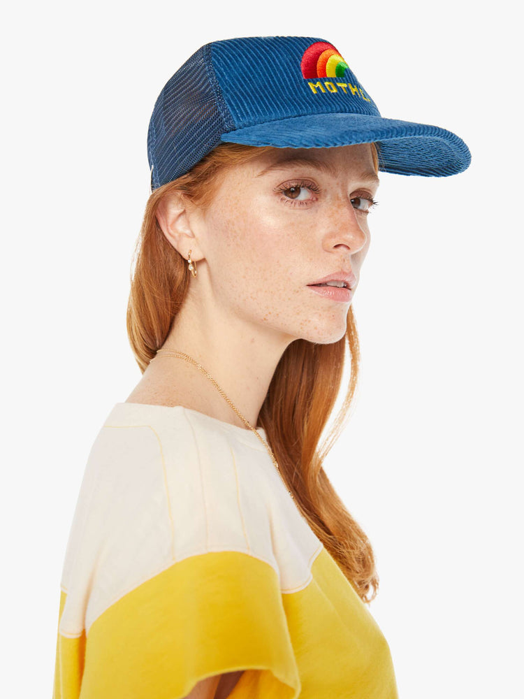 Side view of a woman wearing a blue corduroy trucker hat with a rainbow embroidered.