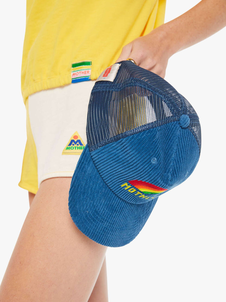 Side view of a woman holding a blue corduroy trucker hat with a rainbow embroidered.