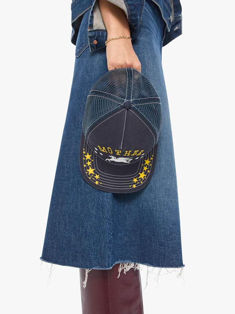Model holding view in a vintage-inspired trucker hat designed in navy with a Pegasus graphic and MOTHER's name in yellow on the front.