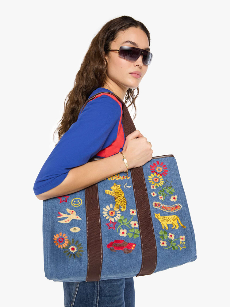 Model holding view of a Tote is designed in mid blue denim with brown top handles, colorful embroidery and MOTHER embroidered across the front in yellow and red.