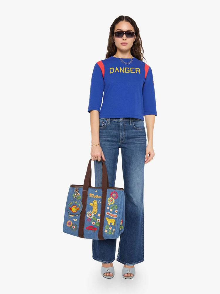 Full body holding view of a Tote is designed in mid blue denim with brown top handles, colorful embroidery and MOTHER embroidered across the front in yellow and red.