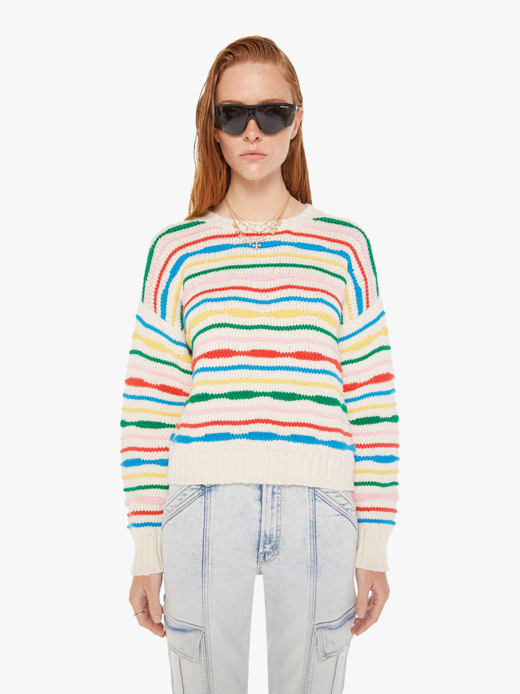 Front view of a woman wearing an off white knit sweater with multi color stripes, featuring dropped shoulders, paired with a light blue acid wash jean.