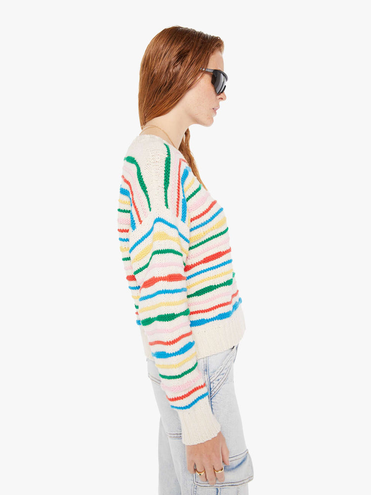 Side view of a woman wearing an off white knit sweater with multi color stripes, featuring dropped shoulders, paired with a light blue acid wash jean.