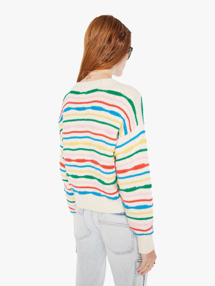 Back view of a woman wearing an off white knit sweater with multi color stripes, featuring dropped shoulders, paired with a light blue acid wash jean.
