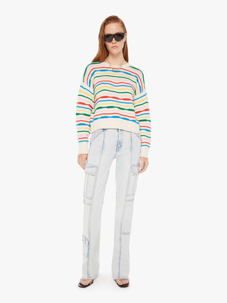 Front full body view of a woman wearing an off white knit sweater with multi color stripes, featuring dropped shoulders, paired with a light blue acid wash jean.