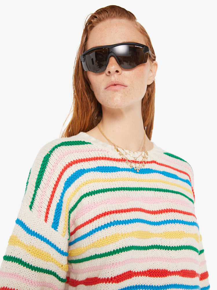 Front close up view of a woman wearing an off white knit sweater with multi color stripes, featuring dropped shoulders.