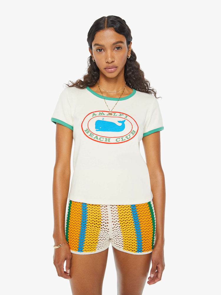 Front view of a woman in an off-white shrunken crewneck tee with extra-short sleeves and a cropped hem designed with a blue whale graphic on the front and teal hems. Paired with crochet shorts.